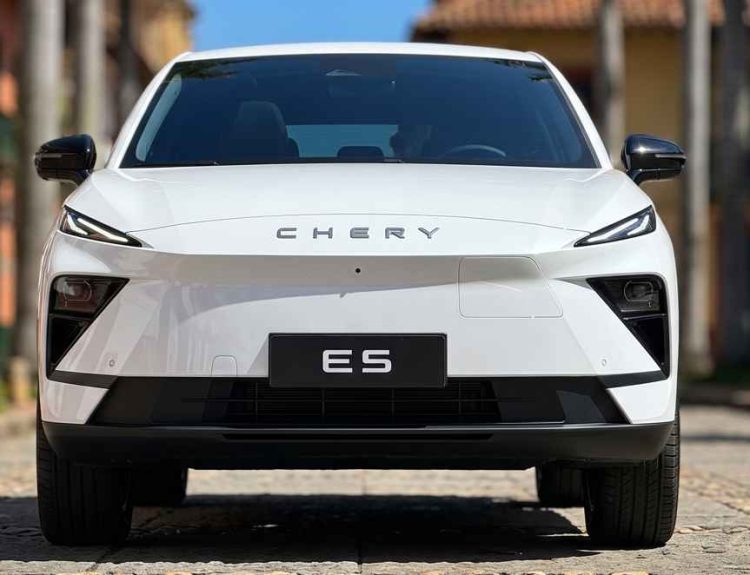 Plano frontal directo de la SUV eléctrica Chery E5 blanca estacionada sobre una calle adoquinada, destacando su diseño frontal cerrado, luces diurnas delgadas y la inscripción Chery en el capó.