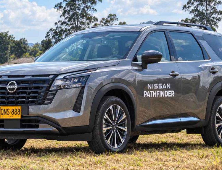 Plano frontal de tres cuartos de la SUV Nissan Pathfinder gris estacionada sobre el pasto junto a un lago, destacando su nueva parrilla ancha con rejillas eslabonadas, rines bitono y faros LED.
