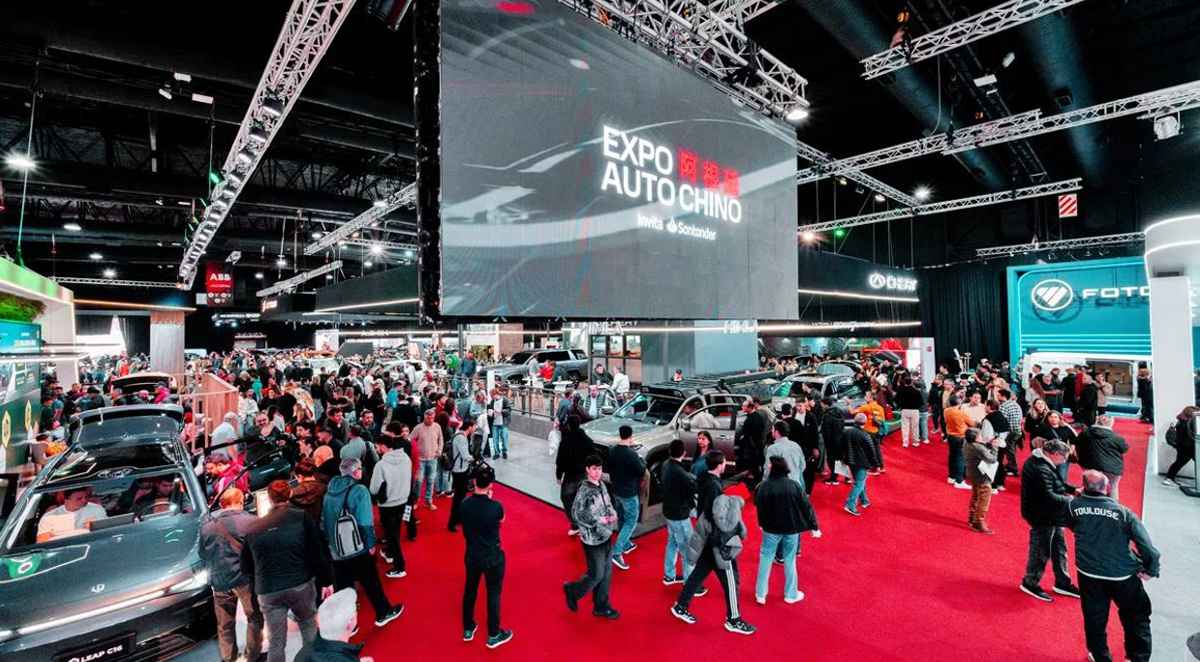 Plano general interior del pabellón principal de la Expo Auto Chino, destacando una multitud de asistentes caminando sobre una alfombra roja bajo la gran pantalla central del evento.