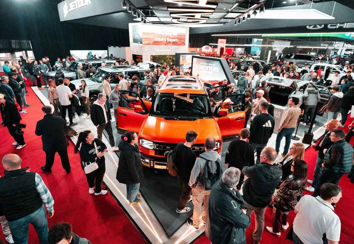 Plano general en picado de la Expo Auto Chino, mostrando a decenas de personas interactuando con una camioneta todoterreno naranja con las puertas abiertas en el centro del recinto. Aparece una Jetour G700.