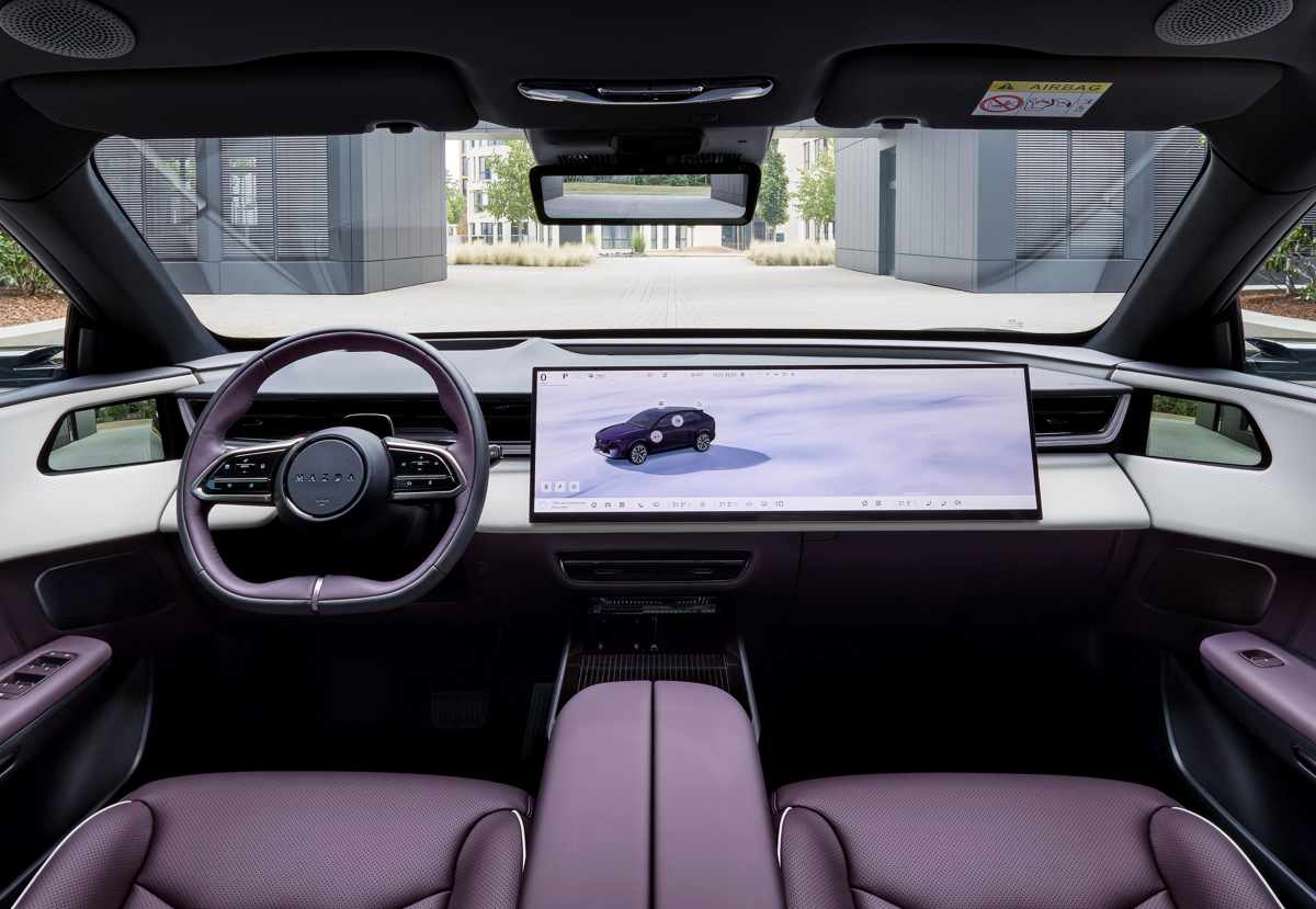 Plano general del tablero interior de la Mazda CX-6e con tapicería en cuero morado y blanco, detallando la gran pantalla central panorámica de 26 pulgadas, el volante ovalado y la ausencia de botones físicos.