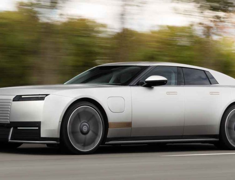 Plano frontal de tres cuartos del gran turismo eléctrico Jaguar Type 01 plateado en movimiento sobre una carretera rodeada de árboles, destacando su capó alargado y la nueva parrilla frontal de líneas horizontales.