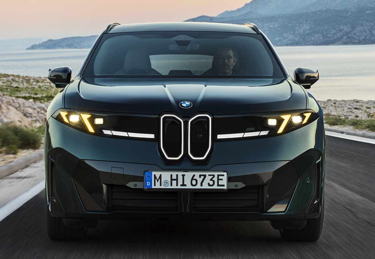Plano frontal directo de la BMW iX3 M60 xDrive verde oscuro circulando por una vía costera, exhibiendo la llamativa firma lumínica de sus luces de competencia M Yellow Lights y los contornos iluminados de la parrilla.