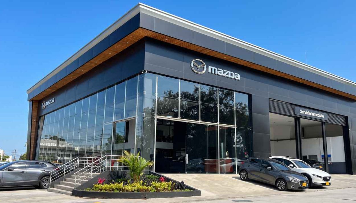Plano frontal de tres cuartos de un moderno concesionario Mazda en Colombia durante un día soleado, destacando su fachada de grandes ventanales, detalles en madera y vehículos parqueados en la entrada.