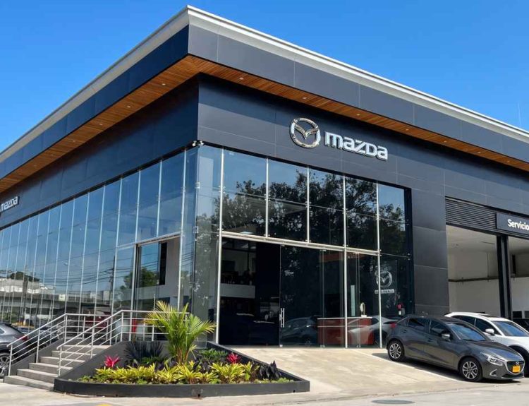 Plano frontal de tres cuartos de un moderno concesionario Mazda en Colombia durante un día soleado, destacando su fachada de grandes ventanales, detalles en madera y vehículos parqueados en la entrada.