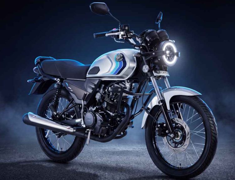 Plano frontal de tres cuartos de la motocicleta AKT NKD 125 color gris níquel sobre fondo oscuro con niebla, destacando su nueva farola redonda con halo LED encendido, tanque de 11,8 litros con franjas azules y rines de radios negros.