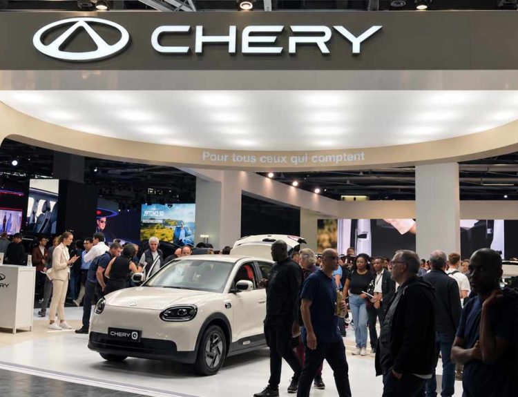 Plano general contrapicado del stand de Chery en el Salón del Automóvil de París 2026, mostrando el aviso iluminado de la marca, visitantes recorriendo el pabellón y el auto eléctrico Chery Q color beige en exhibición.
