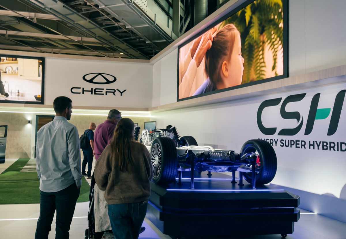 Plano general de la zona tecnológica de Chery en el Salón de París, donde varios visitantes observan la plataforma mecánica Chery Super Hybrid (CSH) con el chasis, suspensión y ruedas al descubierto bajo una pantalla gigante.