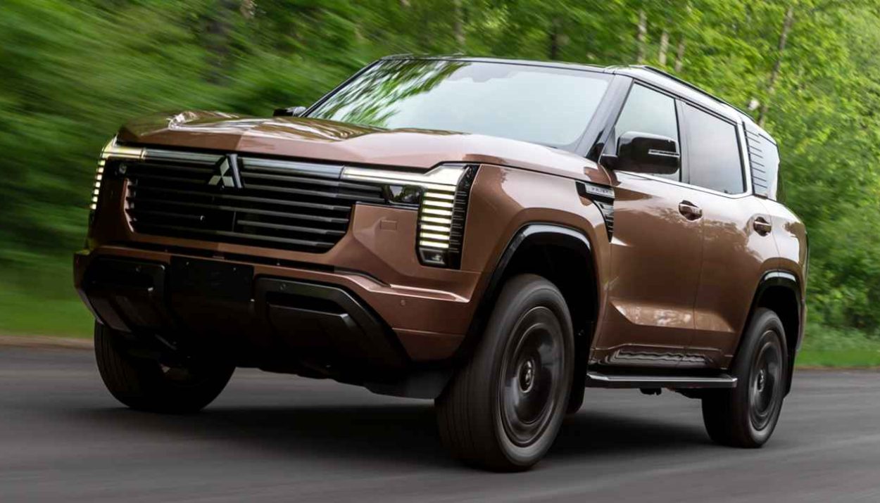 Plano frontal de tres cuartos bajo de la nueva camioneta Mitsubishi Montero 2027 color café bronce rodando en carretera rodeada de árboles, destacando su imponente parrilla negra y faros LED en forma de T.