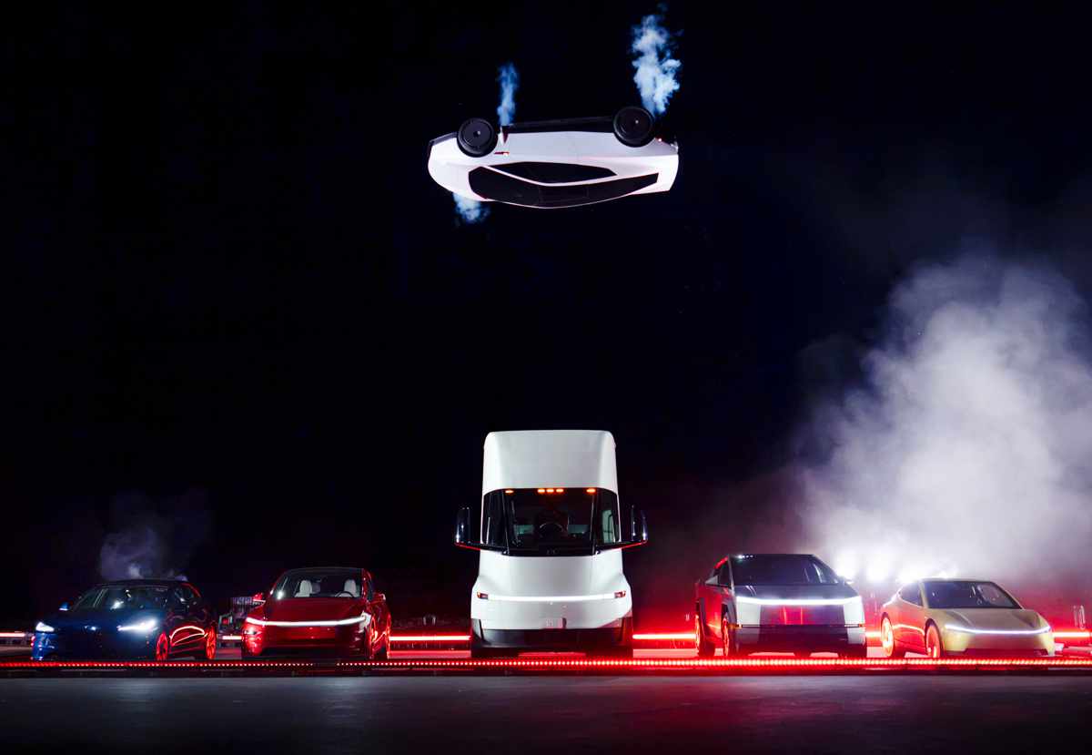 Plano general nocturno del evento en Texas con el Tesla Roadster blanco girando boca abajo en el aire impulsado por cohetes de gas frío, volando sobre una fila de vehículos Tesla que incluye los Model S, Model 3, el camión Tesla Semi, la Cybertruck y el Cybercab.