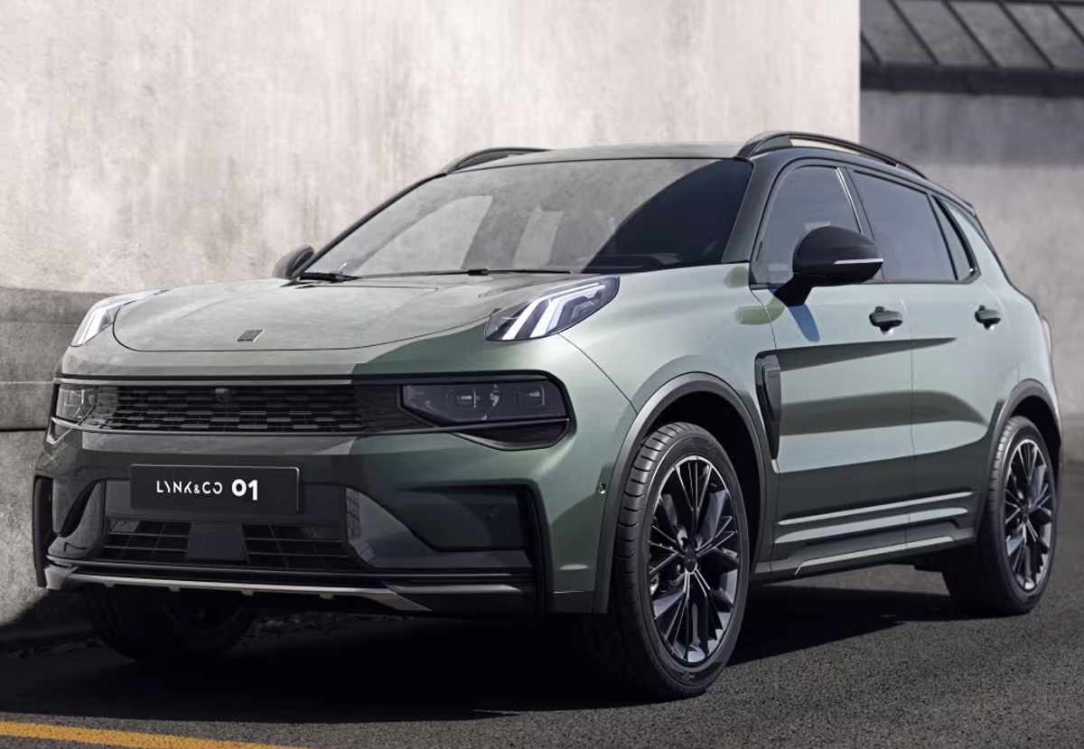 Plano frontal de tres cuartos izquierdo de la SUV híbrida enchufable Lynk & Co 01 color verde oliva mate estacionada junto a un muro de concreto, mostrando su parrilla horizontal negra, luces elevadas y rines deportivos negros.