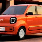 Plano frontal de tres cuartos del miniauto eléctrico FAW Bestune Xiaoma de 5 puertas color naranja frente a una casa moderna al atardecer, destacando sus dos puertas laterales, faros cuadrados redondeados con luz LED y rines blancos carenados.