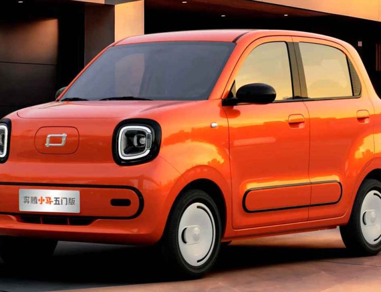 Plano frontal de tres cuartos del miniauto eléctrico FAW Bestune Xiaoma de 5 puertas color naranja frente a una casa moderna al atardecer, destacando sus dos puertas laterales, faros cuadrados redondeados con luz LED y rines blancos carenados.